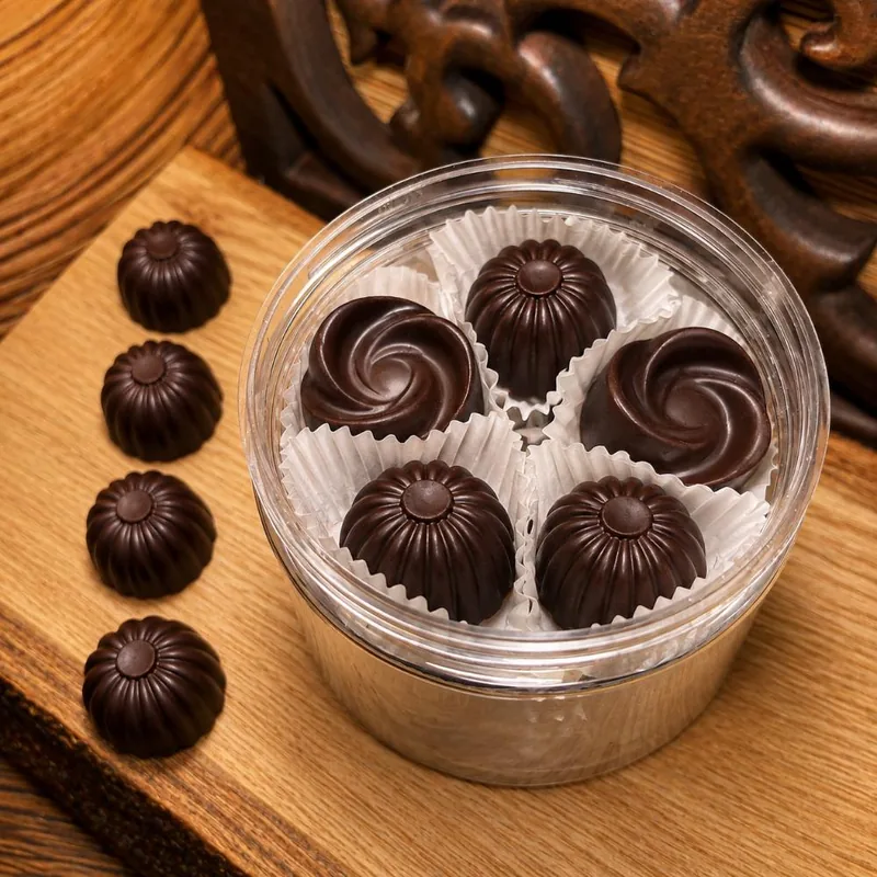 Dark chocolate pralines in white paper cups inside a round tin, with more pralines lined up on a wooden board